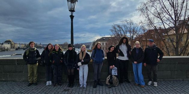 Gruppe von neun Personen steht auf der Karlsbrücke in Prag vor einer Laterne mit Fluss und Gebäuden im Hintergrund bei bewölktem Himmel.