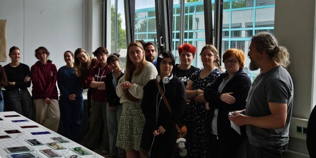 Eine Gruppe von Schülerinnen und Schülern steht in einem Raum mit großen Fenstern und betrachtet einen Tisch, der mit verschiedenen geometrisch geformten Spiegeln bedeckt ist.