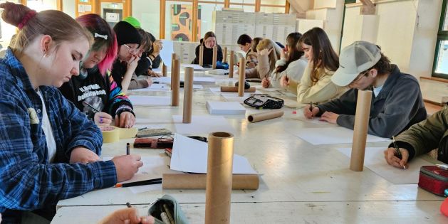Gruppe von Personen sitzt an einem langen Tisch in einem hellen Raum und schreibt oder zeichnet auf Papier