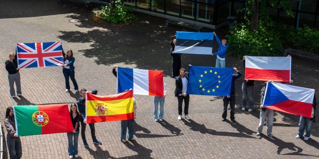 Eine Gruppe von Schülerinnen, Schülern und einer Lehrkraft steht auf einem sonnigen Schulhof und hält lächelnd verschiedene europäische Nationalflaggen sowie die Europaflagge hoch
