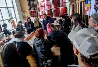 Mehrere Personen sitzen und stehen um einen großen Holztisch in einer Werkstatt mit großen Fenstern und bunten Glasmalereien an der Wand.