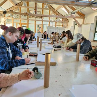 Gruppe von Personen sitzt an einem langen Tisch in einem hellen Raum und schreibt oder zeichnet auf Papier