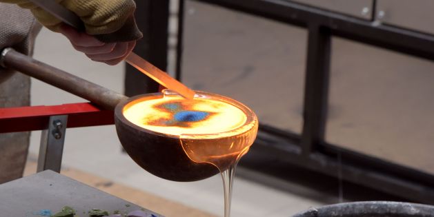 Arbeit am Studioofen beim Internationalen Glassymposium 2025 an der Glasfachschule NRW, geschmolzenes Glas in einer Kelle wird mit blauen Farbpigmenten eingefärbt, blaue Glasstücke auf dem Tisch