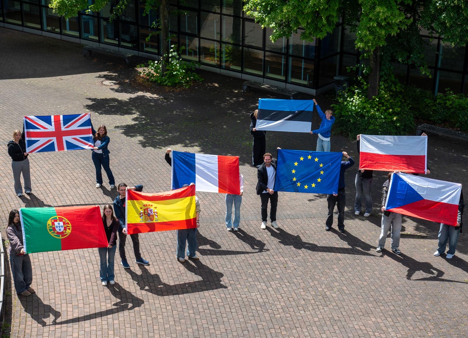 Eine Gruppe von Schülerinnen, Schülern und einer Lehrkraft steht auf einem sonnigen Schulhof und hält lächelnd verschiedene europäische Nationalflaggen sowie die Europaflagge hoch